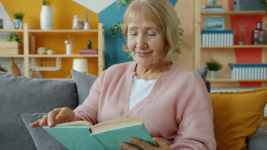 Older woman reading a book on her couch in a bright apartment